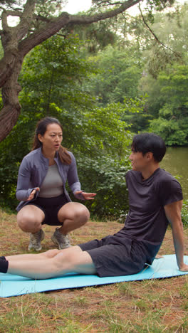 Video-Vertical-De-Una-Mujer-Enseñando-A-Un-Hombre-Que-Viste-Ropa-Deportiva-En-Una-Clase-De-Yoga-Al-Aire-Libre-Rodeada-De-Bosque-Y-Lago-O-Río-Filmado-En-Tiempo-Real